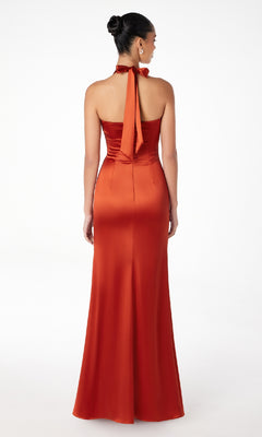 A woman stands facing away in the Bow-Back Long Satin Halter Prom Dress: Astee T2001, a burnt orange, floor-length, backless evening gown with an empire waist and halter neck. The background is plain white.