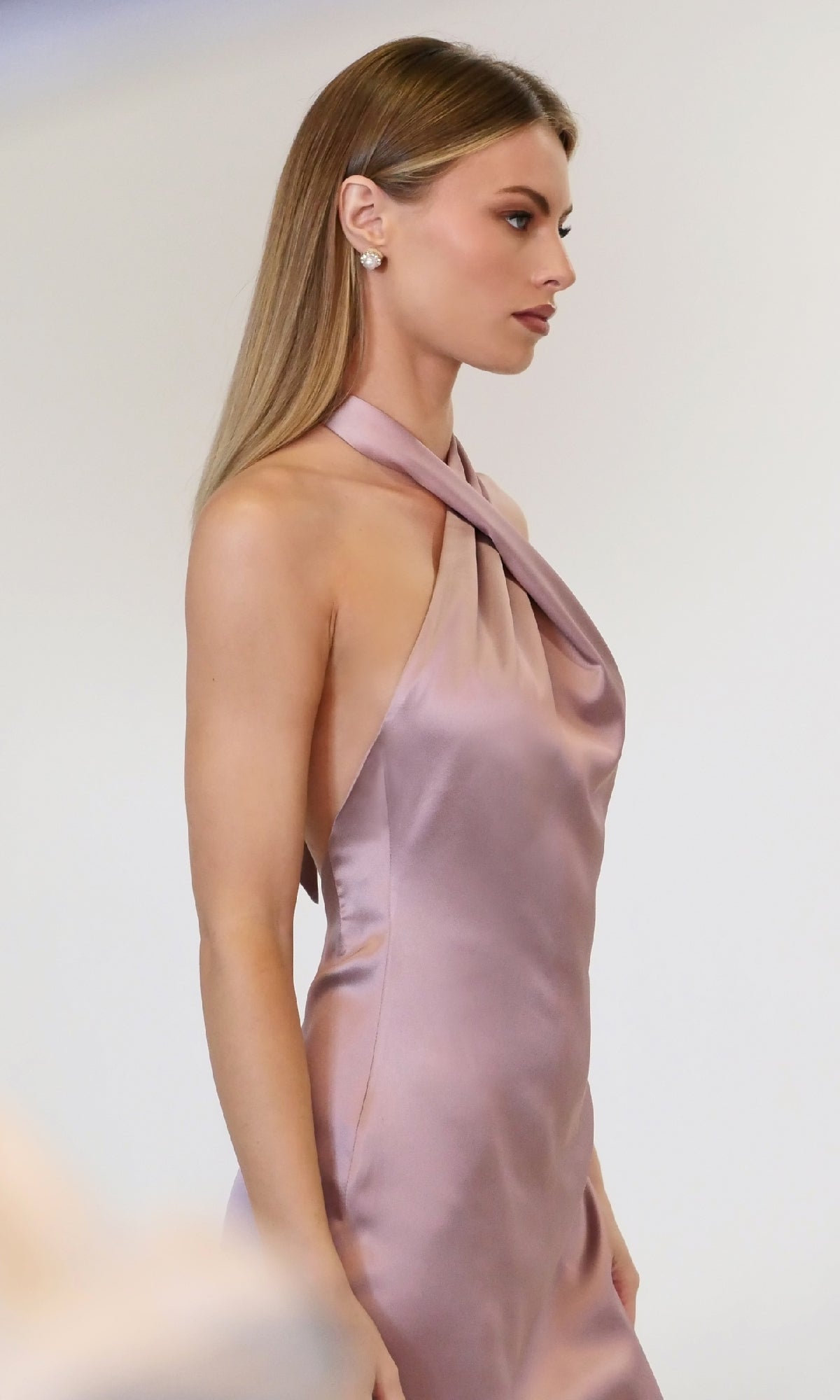 A woman with long straight hair wears the Criss-Cross Halter Long Prom Dress: Astee T2005, a sleeveless light mauve satin gown, along with stud earrings, standing against a plain background.