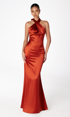 A woman models the Criss-Cross Halter Long Prom Dress: Astee T2005, a fitted, floor-length rust satin gown, against a plain white background.