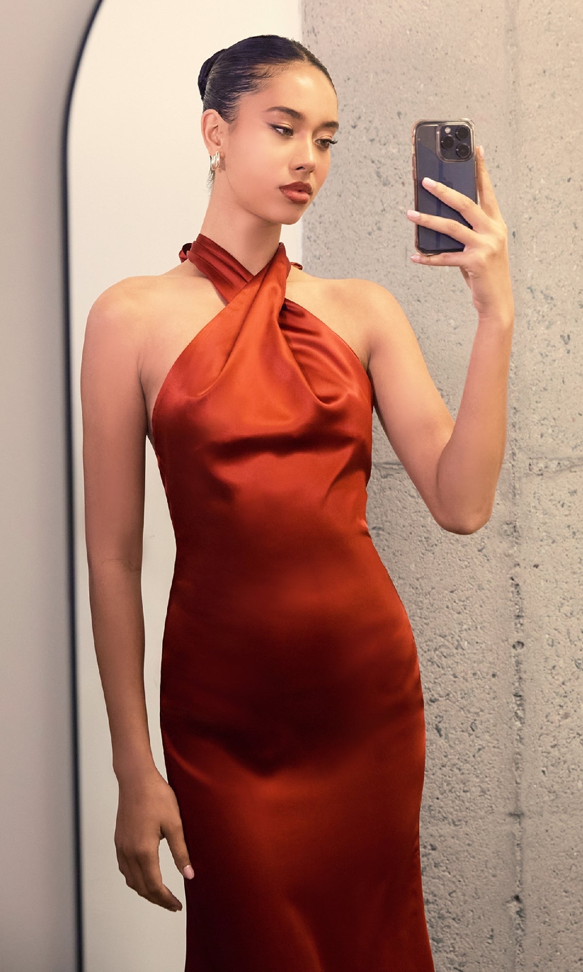 A person in front of a mirror takes a selfie while wearing the Criss-Cross Halter Long Prom Dress: Astee T2005, featuring a shiny, rust-colored satin fabric and an elegant halter neckline.