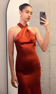 A person in front of a mirror takes a selfie while wearing the Criss-Cross Halter Long Prom Dress: Astee T2005, featuring a shiny, rust-colored satin fabric and an elegant halter neckline.