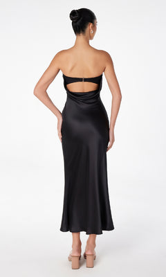 A woman, facing away, wears the Astee T2006 Strapless Long Satin Maxi Prom Dress in black with an open back and cutout detail, paired with nude high heels against a white background.