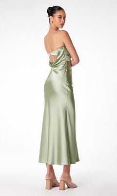 A woman in the Strapless Long Satin Maxi Prom Dress: Astee T2006, light green with an open back and strappy detail, stands facing away and looks over her shoulder. She pairs it with nude heels and a bun hairstyle.