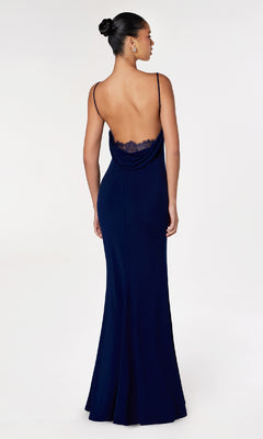 A woman in a navy blue Cowl Open-Back Long Prom Dress: Astee T2008 with spaghetti straps and lace trim stands facing away against a white background, highlighting this affordable evening gown for any special occasion.