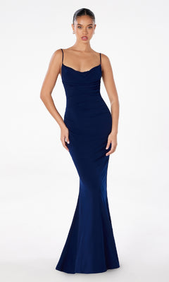 A woman stands against a plain white background wearing the Astee T2008 Cowl Open-Back Long Prom Dress—a fitted, floor-length, dark blue spaghetti-strap formal gown with a slight cowl neckline.