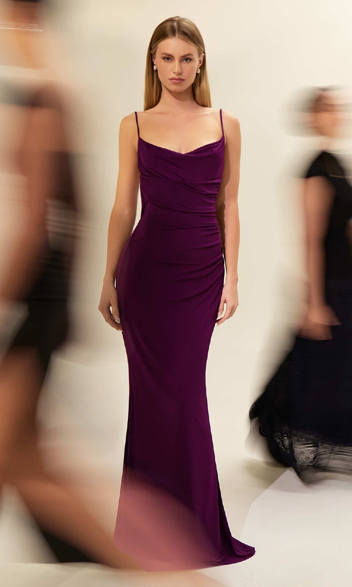 A woman faces forward in the Astee T2008 Cowl Open-Back Long Prom Dress, a fitted, floor-length purple gown with spaghetti straps, while blurred figures move around her against a neutral background.