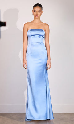 A woman stands against a plain wall wearing the Choker-Sash Strapless Long Prom Dress: Astee T2012, a floor-length, light blue satin gown that exudes elegance.
