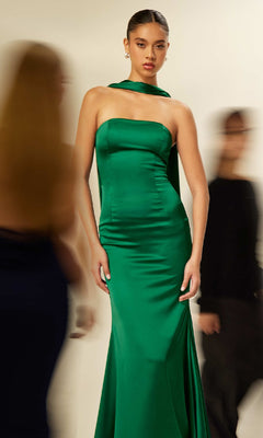 A woman stands wearing the Astee T2012 Choker-Sash Strapless Long Prom Dress, featuring a fitted satin silhouette and scarf neckline, as blurred figures walk past in the background.