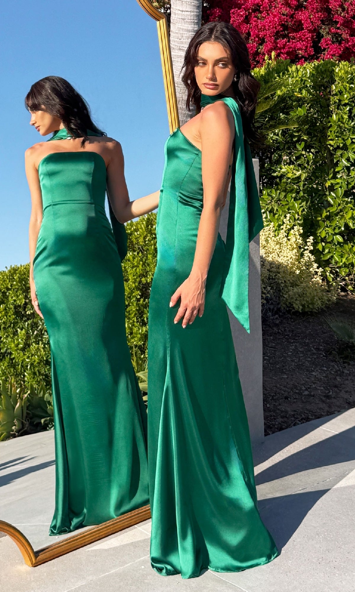 A woman in the Choker-Sash Strapless Long Prom Dress: Astee T2012 poses outdoors by a mirror reflecting her back. Surrounded by lush greenery and pink flowers, she showcases the elegance of this green satin evening gown.