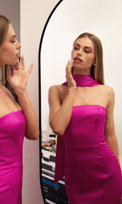A woman in the Choker-Sash Strapless Long Prom Dress: Astee T2012 stands before a mirror, touching her face as she admires her reflection.