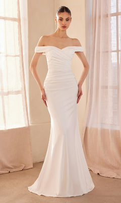 A woman stands indoors in front of tall, sheer-draped windows wearing the Amoris T402—an off-shoulder, long ruched prom dress with a fitted white silhouette and a subtle train.