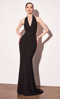 Woman modeling a long black prom dress with a halter cowl neckline standing in front of a white wall.