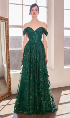 A woman stands indoors wearing the Amoris T607, a long glitter-print prom dress. Facing forward by large windows, she showcases this stunning and affordable emerald green formal gown with off-the-shoulder floral details.