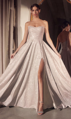 A woman wears the Strapless Long A-Line Glitter Prom Dress Amoris T701—a floor-length silver gown with a thigh-high slit and silver heels—standing indoors by a curtain and mirror.