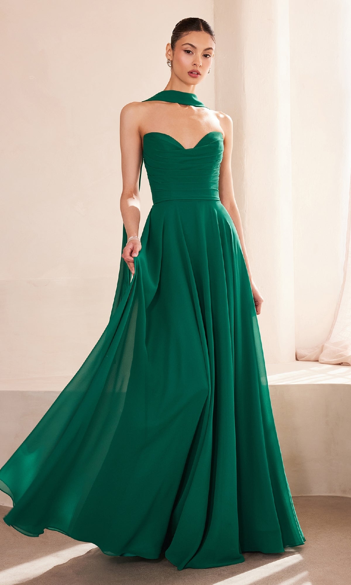 A woman stands indoors in the Amoris T703 Strapless Long Chiffon Prom Dress, a floor-length emerald green gown with a sweetheart neckline, corset bodice, and matching neck scarf.