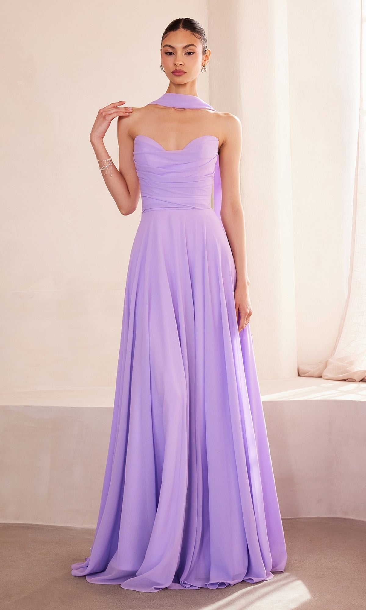 A woman poses indoors in the affordable Strapless Long Chiffon Prom Dress: Amoris T703—an elegant, floor-length lavender gown with a flowing skirt, corset bodice, and a matching scarf around her neck.