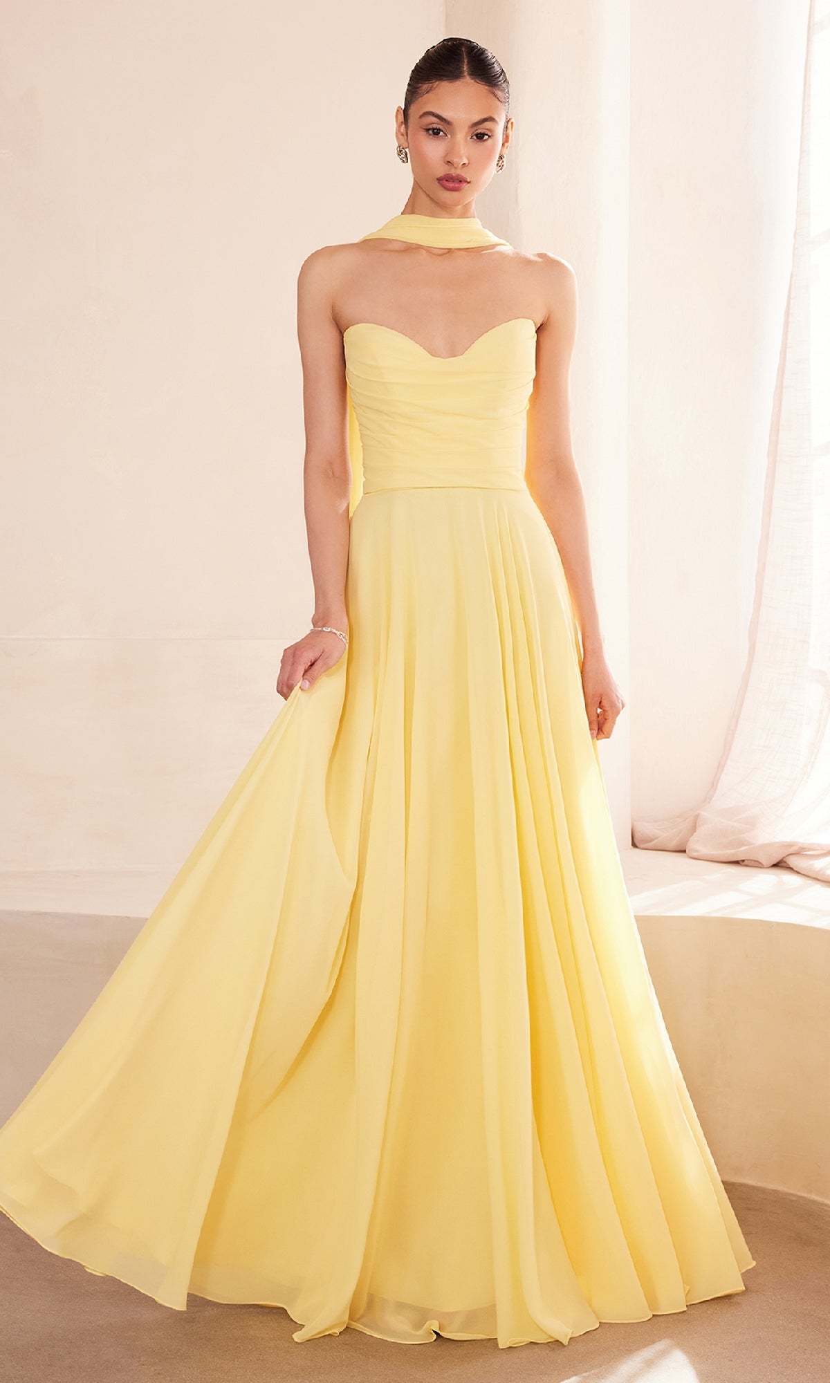 A woman in the Strapless Long Chiffon Prom Dress: Amoris T703 in light yellow, paired with a sheer scarf, stands indoors near a softly lit window.
