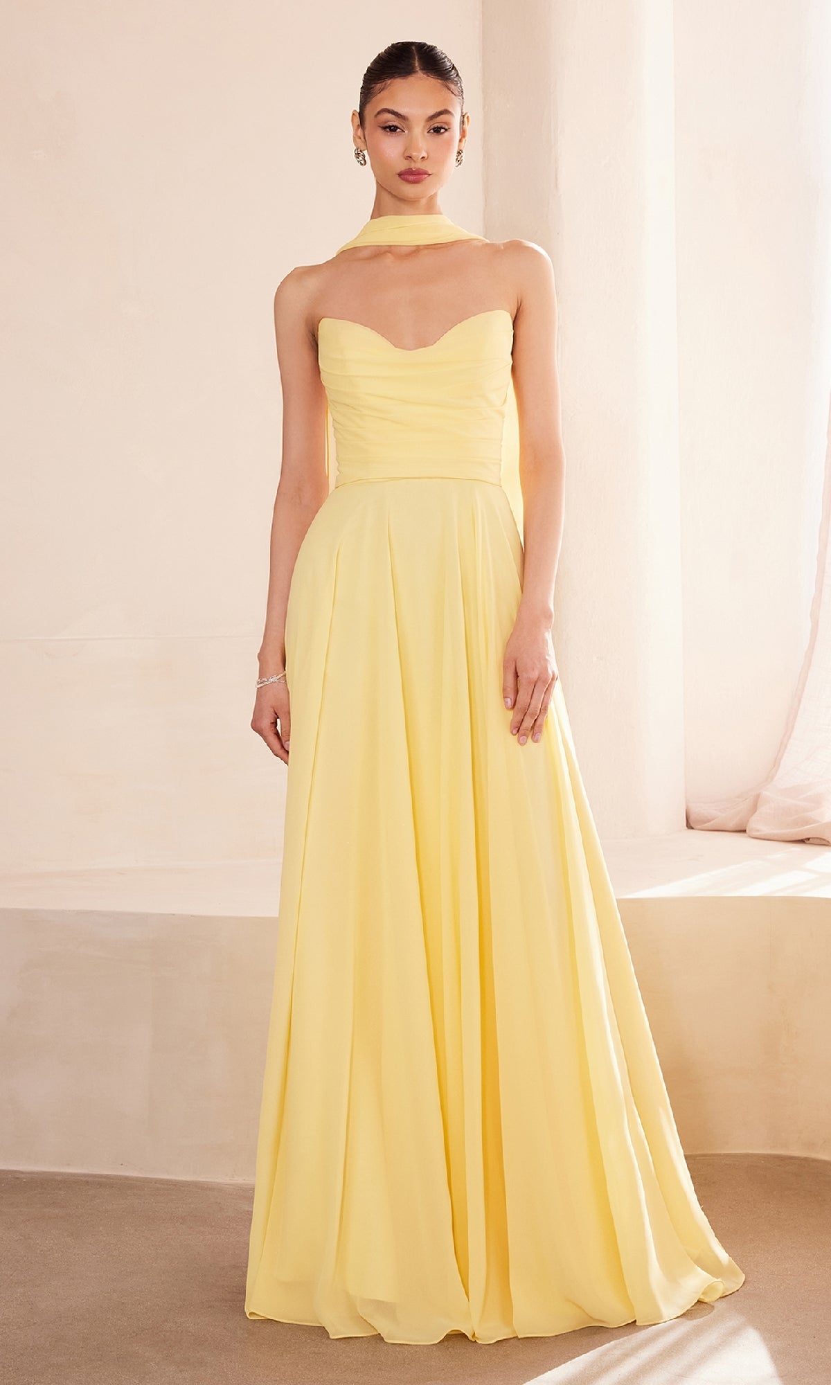 A woman stands indoors wearing the Strapless Long Chiffon Prom Dress: Amoris T703, a yellow, floor-length gown with a matching neck scarf.