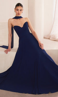 A woman sits elegantly against a light-colored wall, wearing the Strapless Long Chiffon Prom Dress: Amoris T703—a navy blue gown with a corset bodice, sweetheart neckline, and matching scarf.