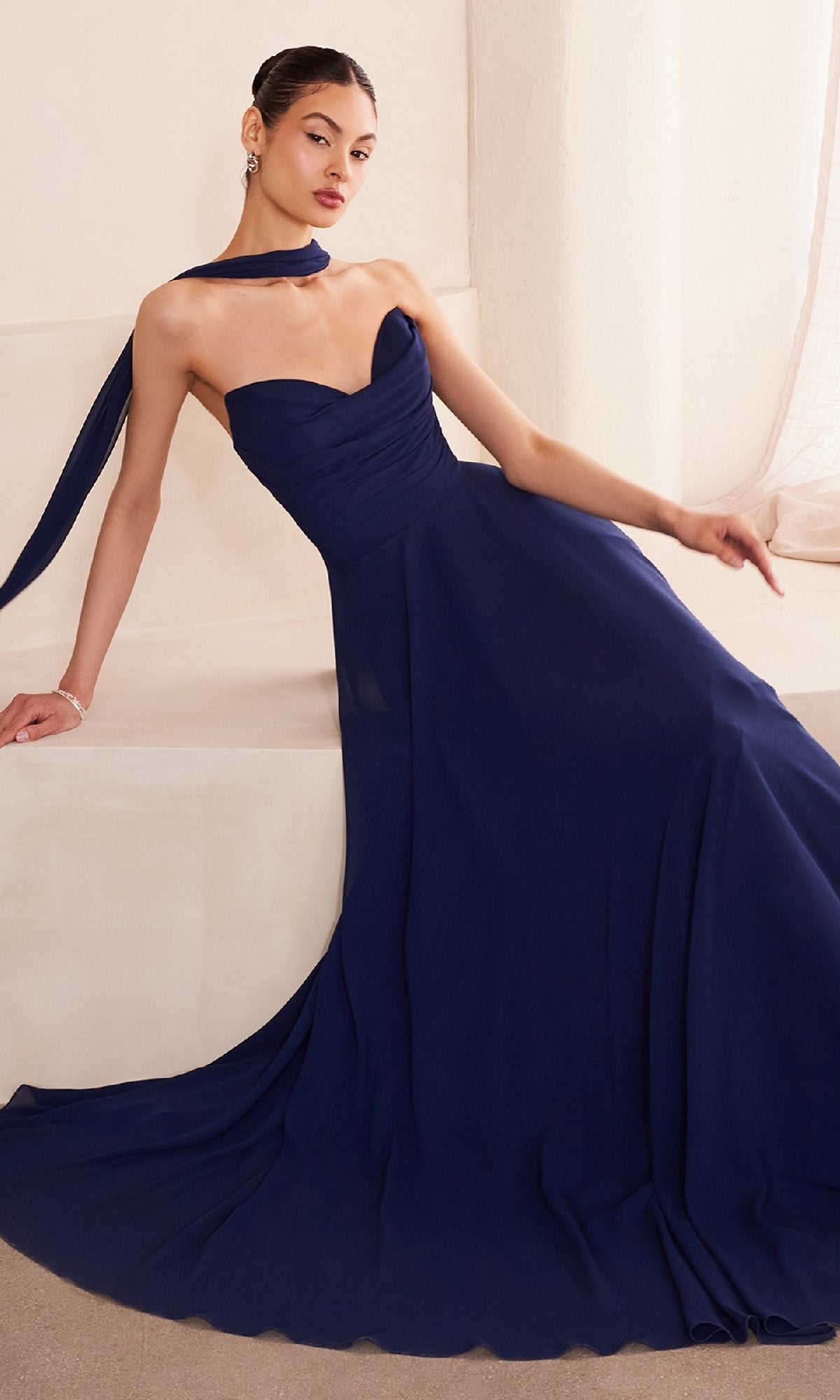 A woman in the affordable Strapless Long Chiffon Prom Dress: Amoris T703 with a matching scarf sits elegantly on a white ledge in a softly lit room.
