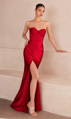 A woman stands indoors wearing the Amoris T708 Strapless Faux-Wrap Long Prom Gown, featuring red satin with a structured bodice and thigh-high slit, paired with clear high-heeled sandals.