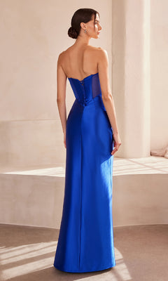 A woman stands indoors, facing away, in the Strapless Faux-Wrap Long Prom Gown: Amoris T708—an elegant royal blue dress with a fitted bodice and floor-length skirt, perfect for formal occasions.