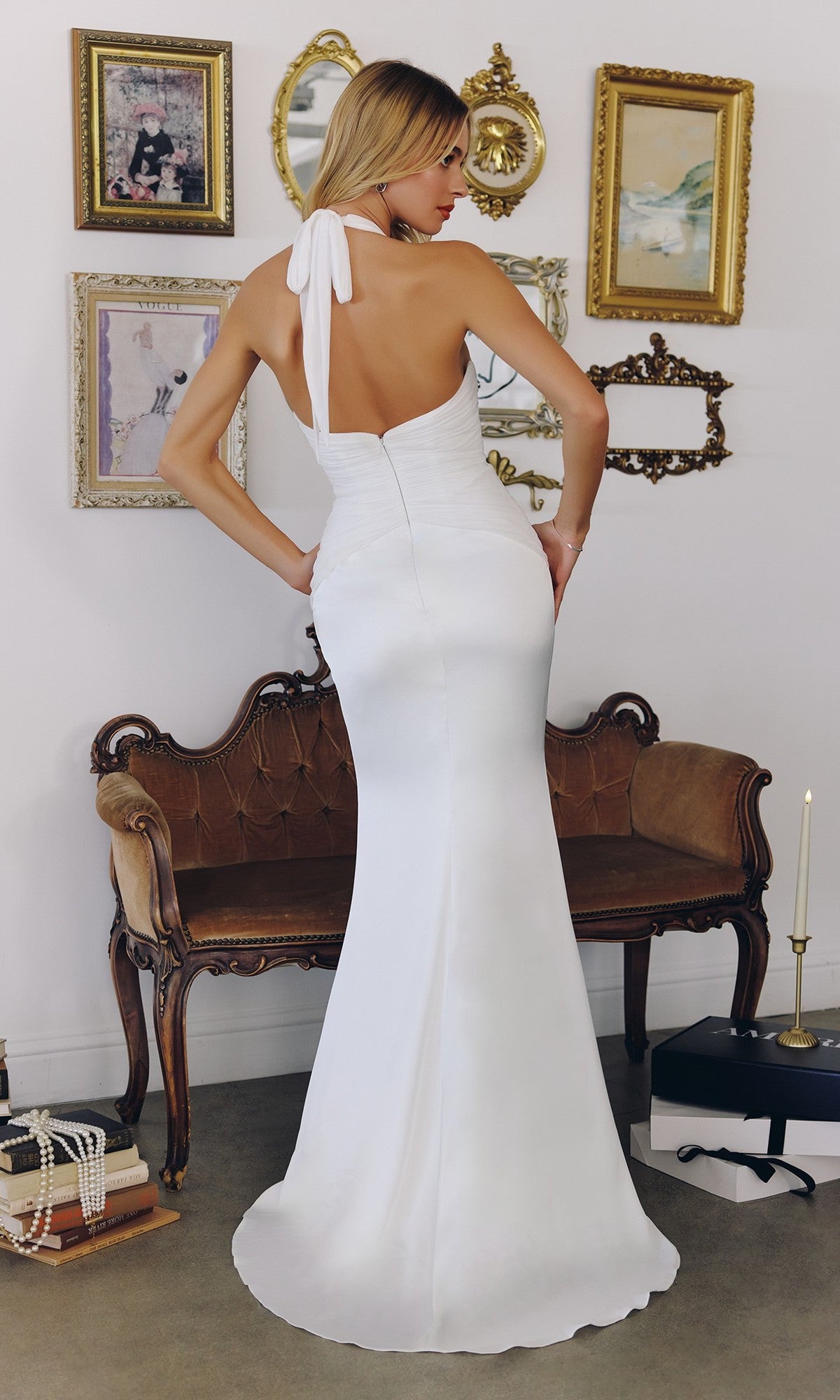A woman in the Amoris T720W long prom dress, featuring an off-white halter v-neckline, stands with her back to the camera in a vintage-style room with framed art and a brown settee.