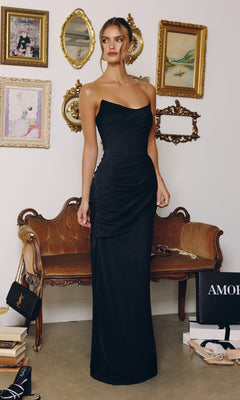 A woman in the black Amoris T726 long prom dress, featuring a strapless chiffon design and lace-up back, stands before a vintage-style sofa with framed art and decorative mirrors on the wall behind her.