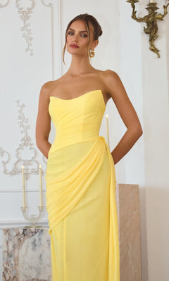 A woman wearing the Amoris T726 Long Prom Dress, featuring a strapless chiffon design with draped details and a lace-up back, stands indoors by a candelabra and marble surface.