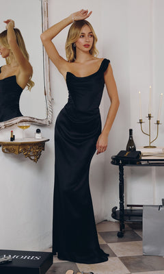 A woman wearing the black Amoris T727 long prom dress stands in a room with a mirror, wine glass, candle holder, and shopping bags in the background.