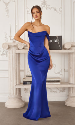 A woman stands indoors wearing the Amoris T727 fitted long prom dress in royal satin with an off-the-shoulder neckline, posed before a white mantel decorated with books and candles.