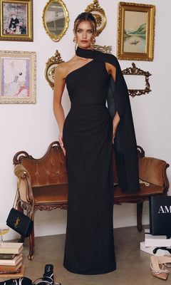 A woman wearing the Amoris T730 long black prom dress with a ruched bodice stands in a stylish room featuring vintage decor like a velvet sofa, framed art, gold mirrors, and designer shopping bags.