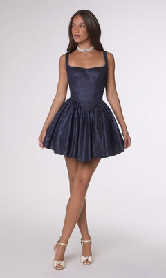 A woman wearing the Amoris T738 short homecoming dress in dark blue shimmer stretch fabric, paired with white heels, stands against a plain light background.