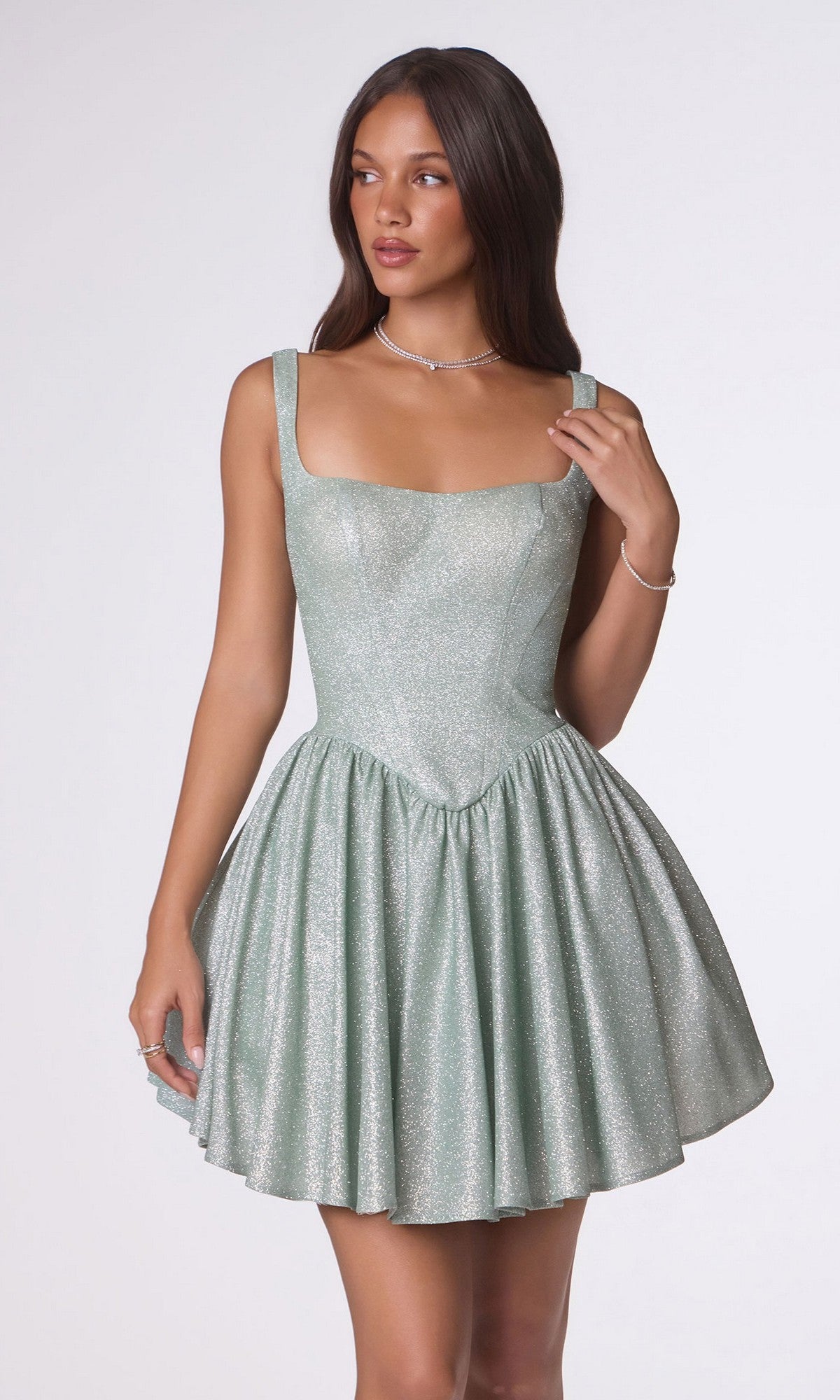 A woman wears the Amoris T738 short homecoming dress in sage, featuring a sleeveless fitted bodice and flared skirt crafted from shimmering stretch fabric, standing against a plain light background.