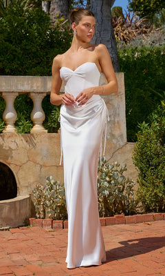 A woman wearing the Amoris T747W long prom dress in off white, featuring a flattering corset bodice and strapless neckline, stands on a brick patio with greenery and a stone balustrade in the background.