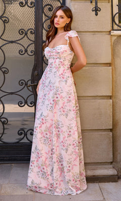 A woman wearing the Long Prom Dress: Amoris T749—a light pink floral gown—stands by an ornate black wrought iron gate and stone wall, looking at the camera.