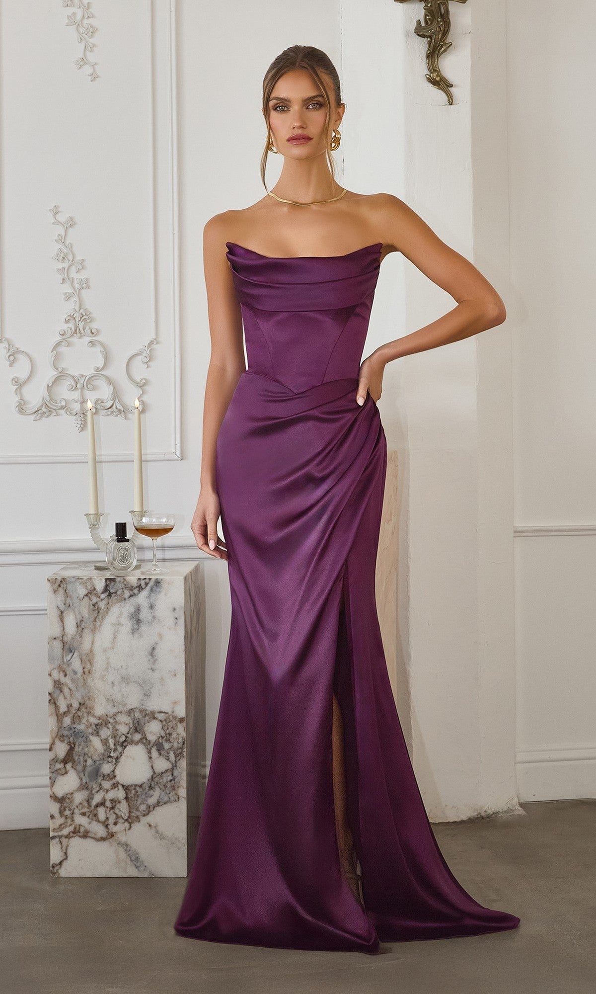 A woman poses indoors in the Amoris T820 Long Prom Dress in eggplant satin, featuring an off-the-shoulder design and a thigh-high slit. She stands beside a marble table with candles and a glass of drink.