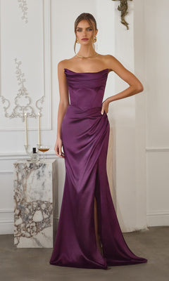 A woman poses indoors in the Amoris T820 Long Prom Dress in eggplant satin, featuring an off-the-shoulder design and a thigh-high slit. She stands beside a marble table with candles and a glass of drink.