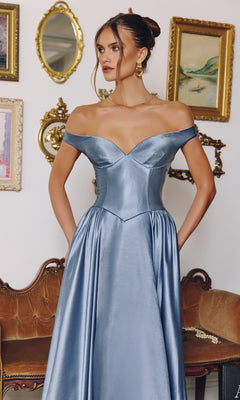 A woman stands before a vintage-style wall and brown sofa, wearing the Amoris T821 long prom dress—an off-the-shoulder, satin blue gown with pockets and an elegant A-line silhouette.
