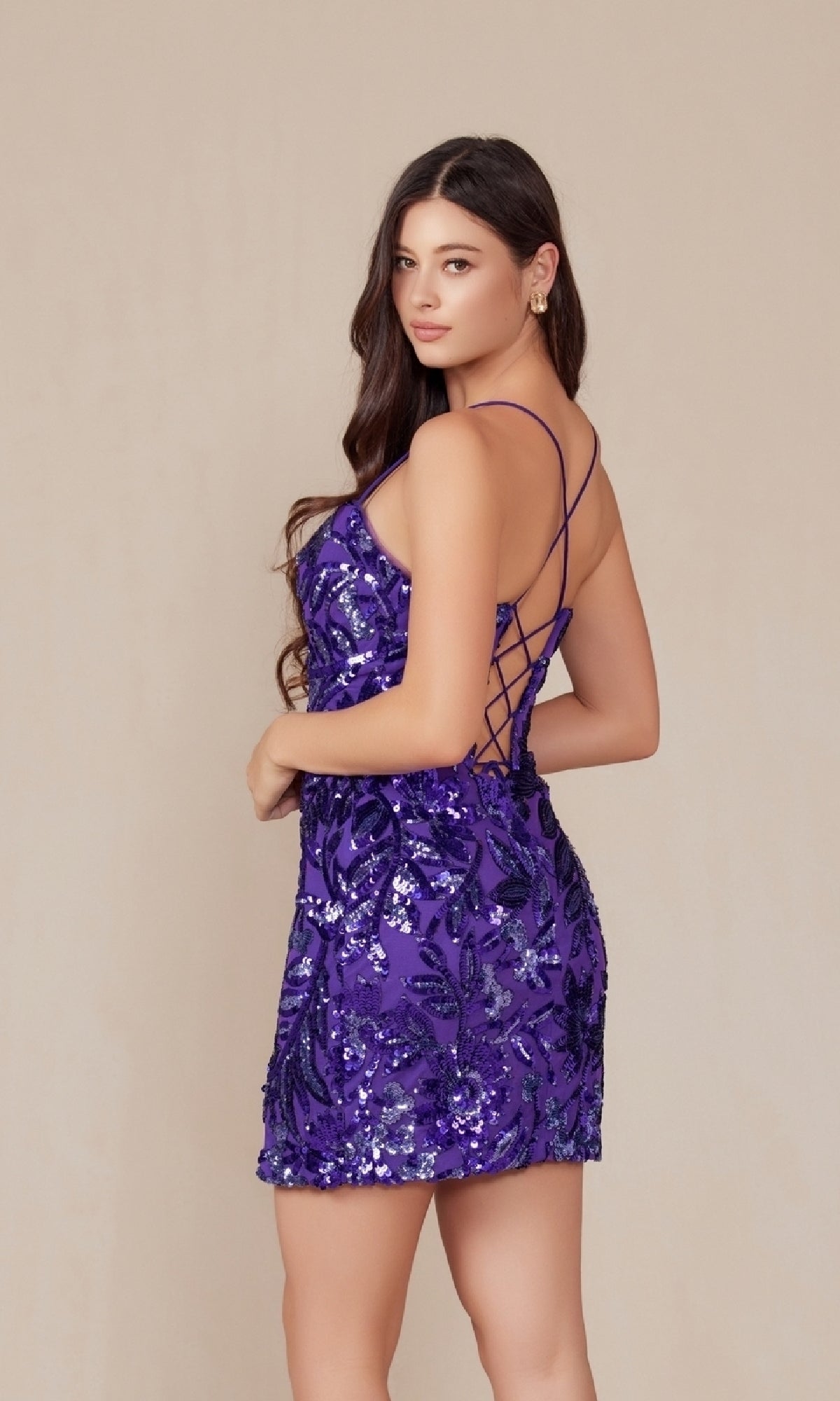 A woman poses in the Lace-Up Short Sequin-Print Homecoming Dress T864—a sparkly, sleeveless purple mini dress with crisscross straps and an open back—standing against a plain beige background.