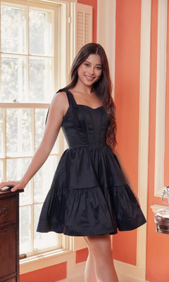 A woman stands indoors by a window, smiling at the camera in the Shoulder-Tie Short Lace-Up A-Line Hoco Dress T869—a stylish sleeveless black dress with a fitted bodice and flared skirt, perfect for any semi-formal party.