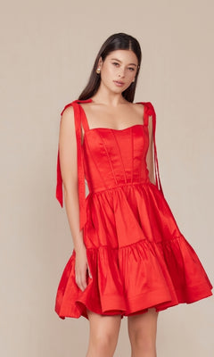 A woman poses against a plain beige background, wearing the Shoulder-Tie Short Lace-Up A-Line Hoco Dress T869—a sleeveless, tiered a-line dress with ribbon tie straps.