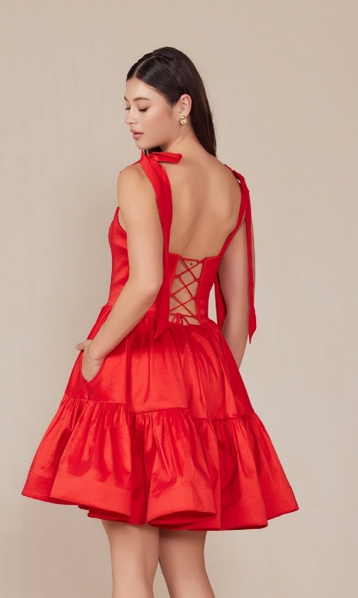 Woman wears the Shoulder-Tie Short Lace-Up A-Line Hoco Dress T869—a red, sleeveless dress with bow straps and a tiered skirt—against a beige background. This stylish short A-line design is perfect for any semi-formal party.