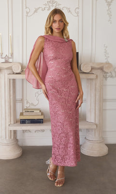A woman models the Amoris T906 Long Prom Dress—sleeveless and floor-length with sequin-lace and a sheer cape—standing indoors before a white mantel decorated with books and candles.