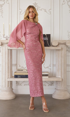 A woman stands indoors in a Dusty Rose Long Prom Dress: Amoris T906 with sequin-lace details and matching chiffon cape overlay, posing before a decorated white mantel lined with books.