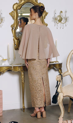 A woman in the Amoris T906 Long Prom Dress, featuring sequin lace and a sheer chiffon cape overlay, stands before a vintage vanity adorned with candles and ornate decor, admiring her reflection.