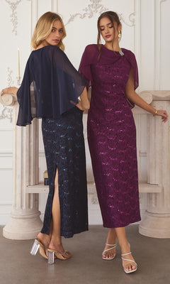 Two women in formal wear: one in the Amoris T906 long prom dress, a navy blue sequin-lace gown with sheer cape and side slit; the other wears a purple dress with matching chiffon cape overlay, both standing in an elegant room with classic columns.
