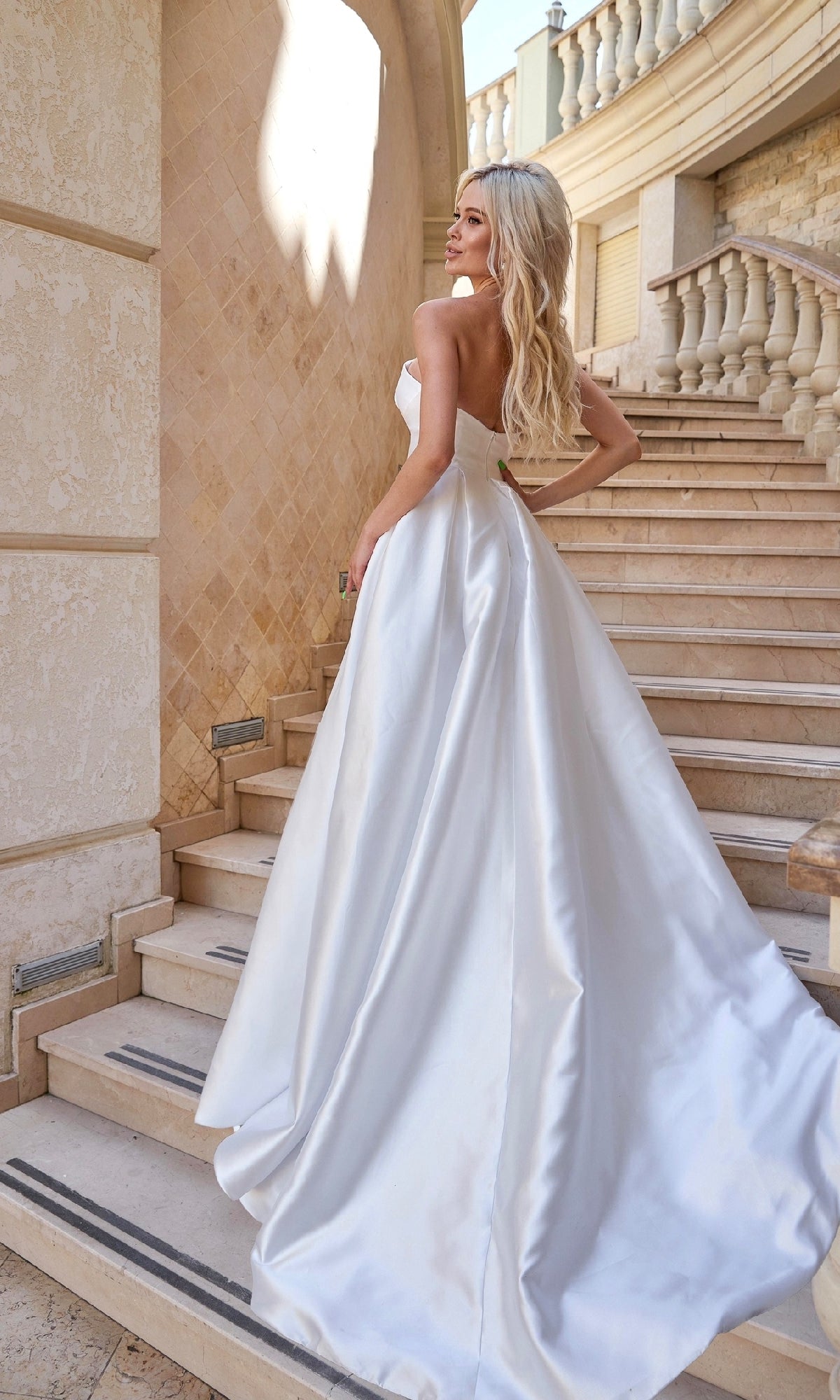 A woman wearing the Strapless Off-White Bridal Gown: Tina Holly TA611W stands on a stone staircase, facing away and looking over her shoulder.
