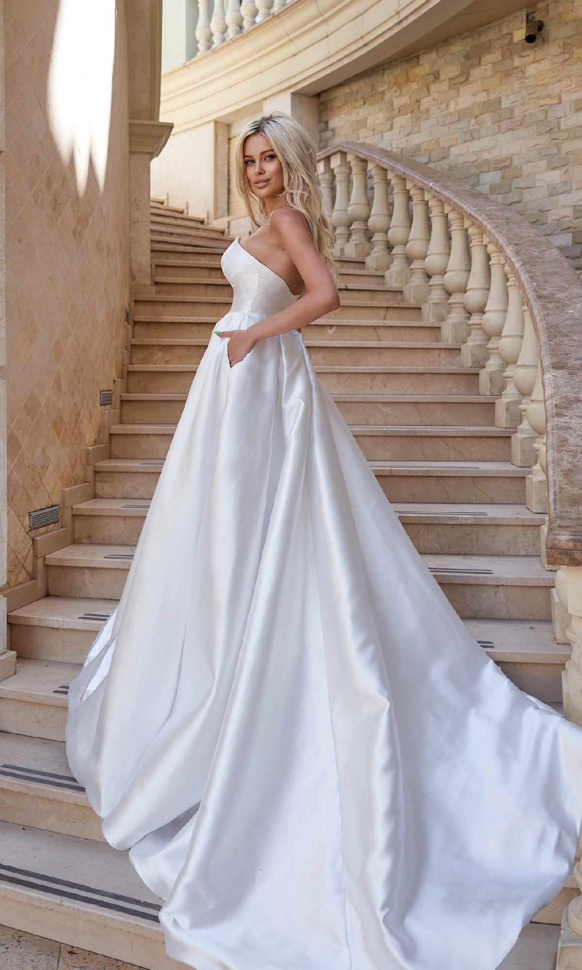 A woman wearing the Strapless Off-White Bridal Gown: Tina Holly TA611W stands on a curved stone staircase, with a brick wall and balustrade in the background.