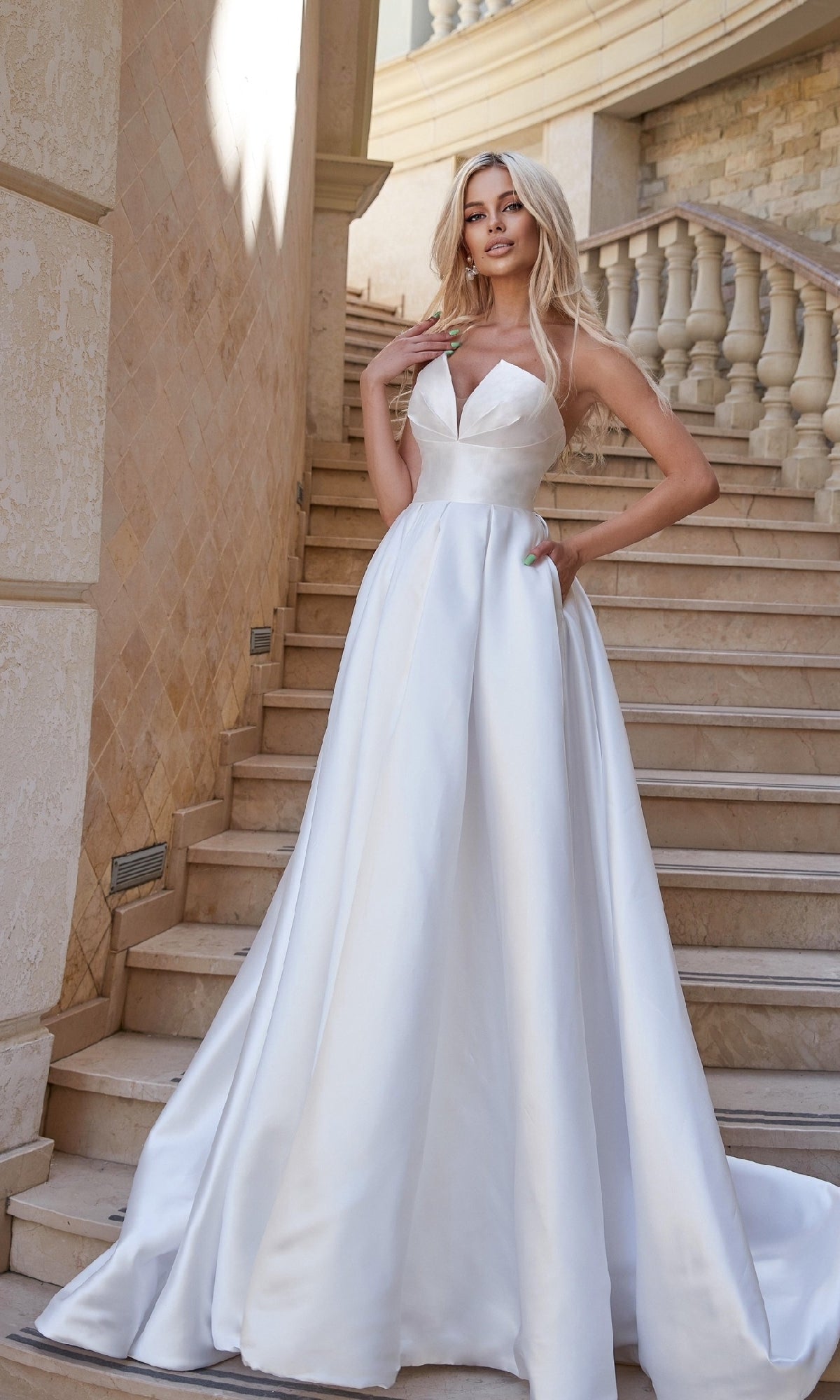 A woman stands on a stone staircase beside a balustrade, wearing the Tina Holly TA611W strapless off-white bridal gown with a fitted bodice and full skirt.